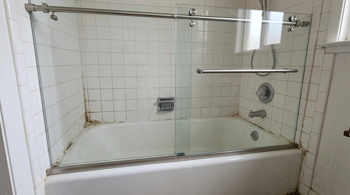 After: The same bathroom and alcove with the old framed door replaced by a crystal-clear frameless sliding glass door with brushed nickel hardware, same tile, same lighting