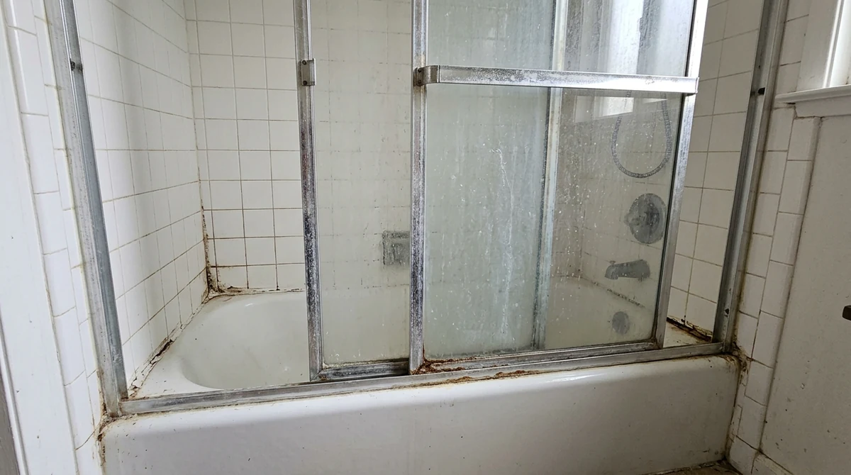 Before: An older bathroom shower alcove with a dated aluminum-framed sliding glass door, cloudy water-stained glass and a corroded bottom track, white tile walls, a tub below, bright light