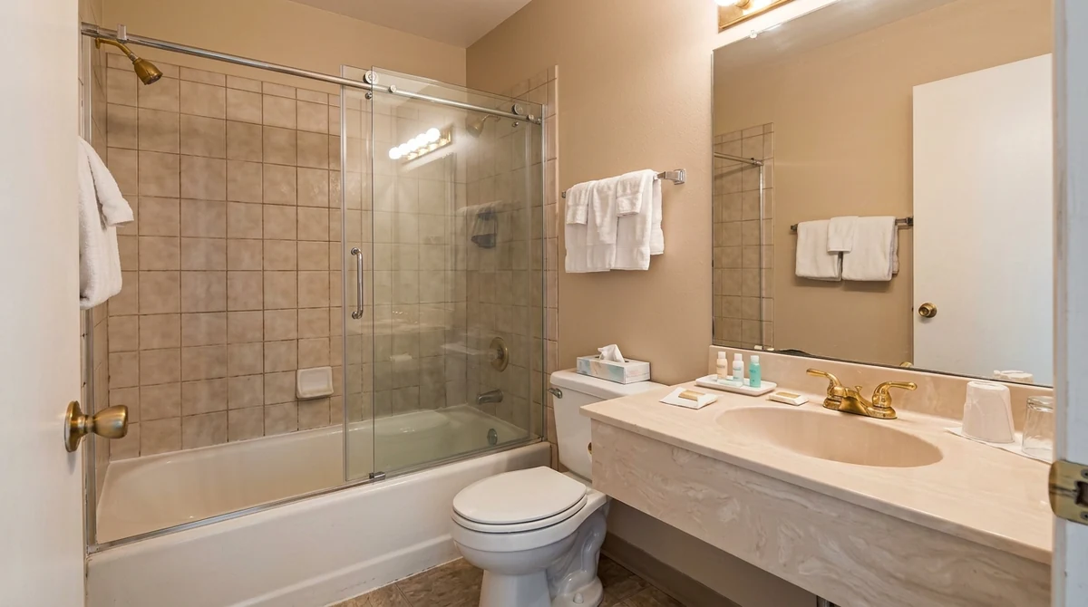 After: The same hotel bathroom with a sleek frameless clear glass shower enclosure with polished chrome hardware, same tile and vanity, same lighting
