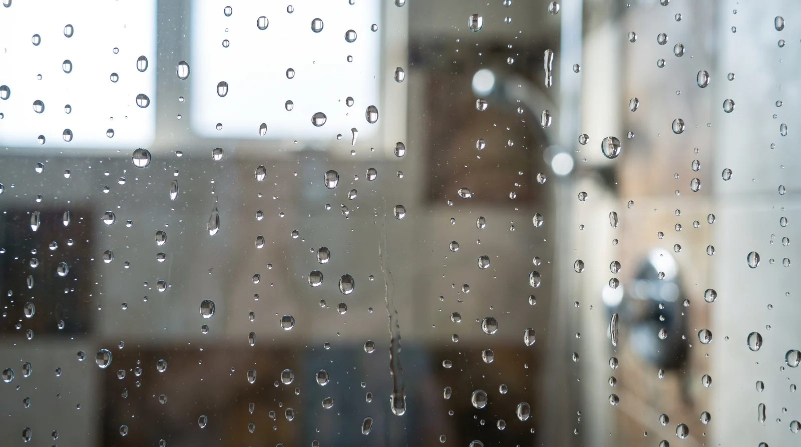 A macro of crisp water droplets beading on perfectly clear shower glass with a soft out-of-focus tiled shower behind, bright backlight, sharp droplets