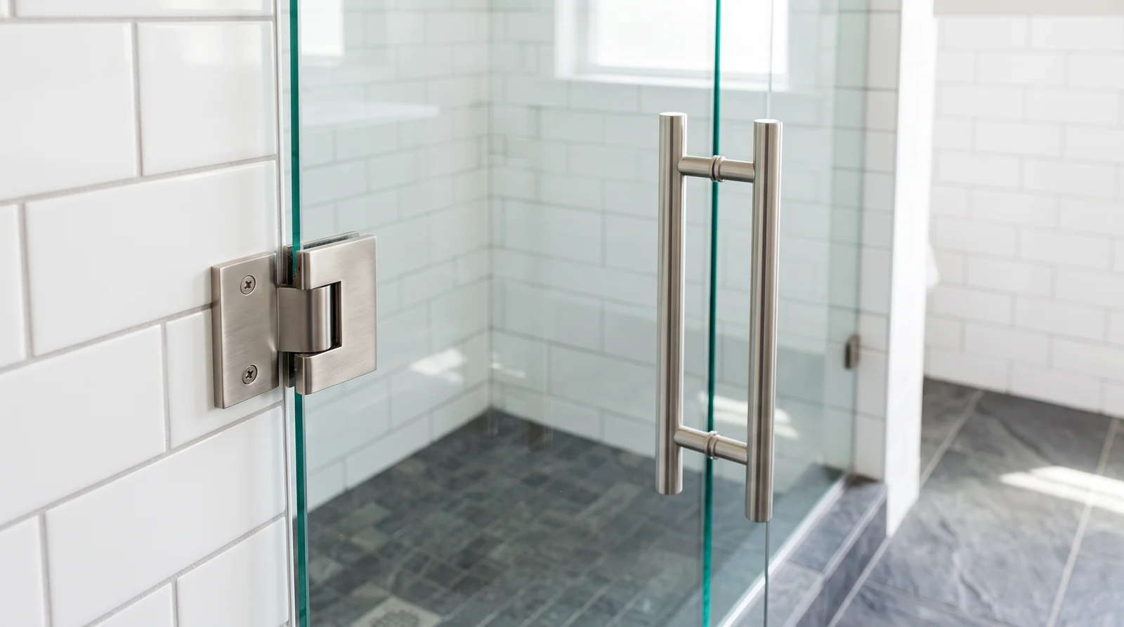 A close three-quarter view of a frameless clear glass shower door with brushed nickel hinges and a sleek bar handle in a bright modern bathroom, white subway tile and a gray stone floor, soft daylight, shallow depth of field on the hardware