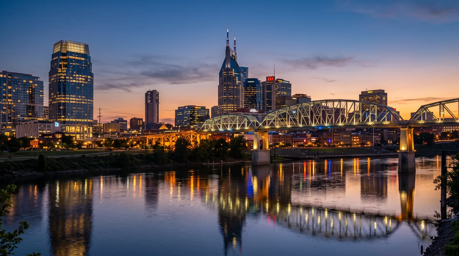 The Nashville skyline glowing at blue-hour dusk beyond the Cumberland River with a calm reflection, a warm sunset gradient in the sky and the lit pedestrian bridge in the foreground