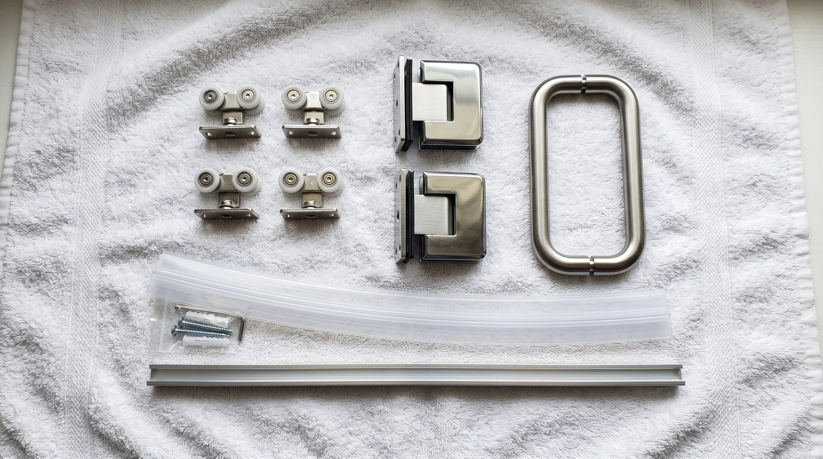Rollers, Hinges and Hardware | Music City Shower Doors A flat-lay of new shower door rollers, hinges, a handle, a door sweep and a bottom track on a white towel, sharp detail, soft light