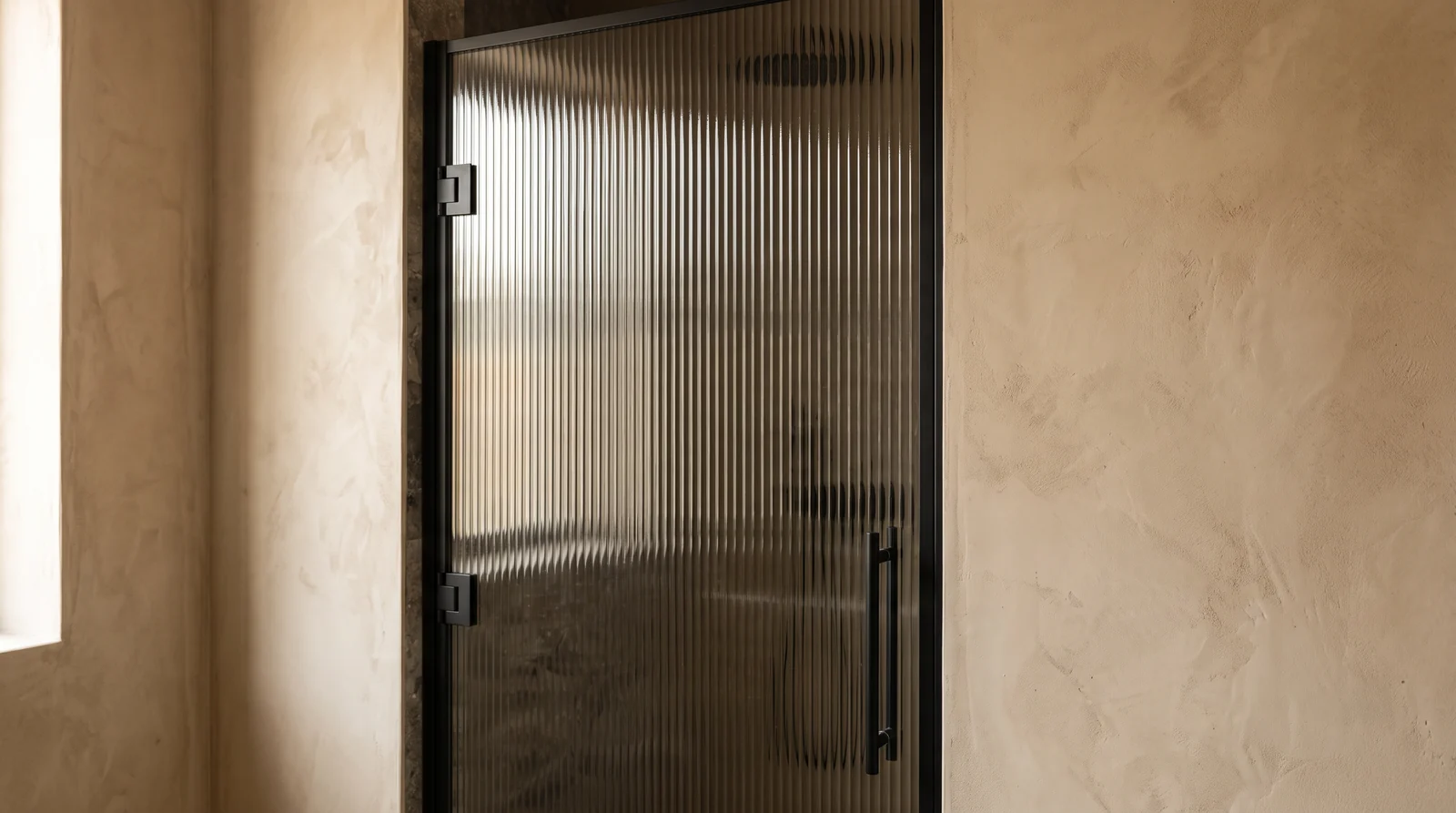 A shower door with vertical reeded textured glass catching soft light with beautiful distortion, matte black hardware and warm plaster walls