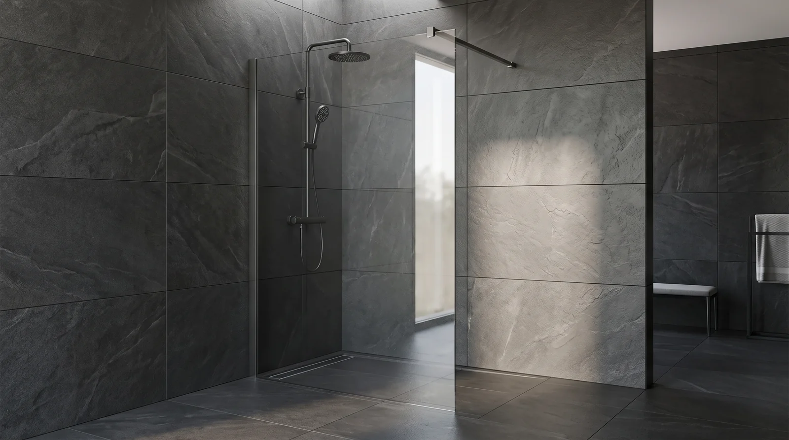 A walk-in shower with a single clear fixed glass panel and a stabilizing bar, no door, a linear drain and large-format dark tile, minimalist modern bathroom, soft light